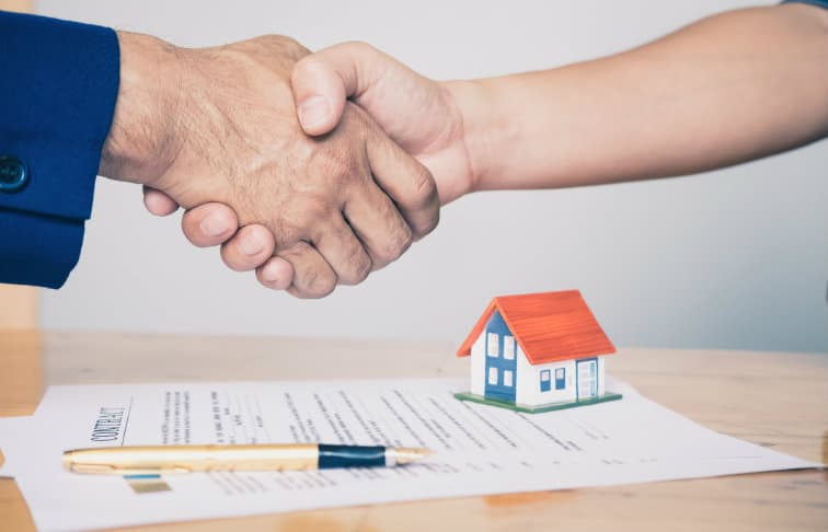 real estate agency - two people shaking hands over real estate contract with miniature house on top of it